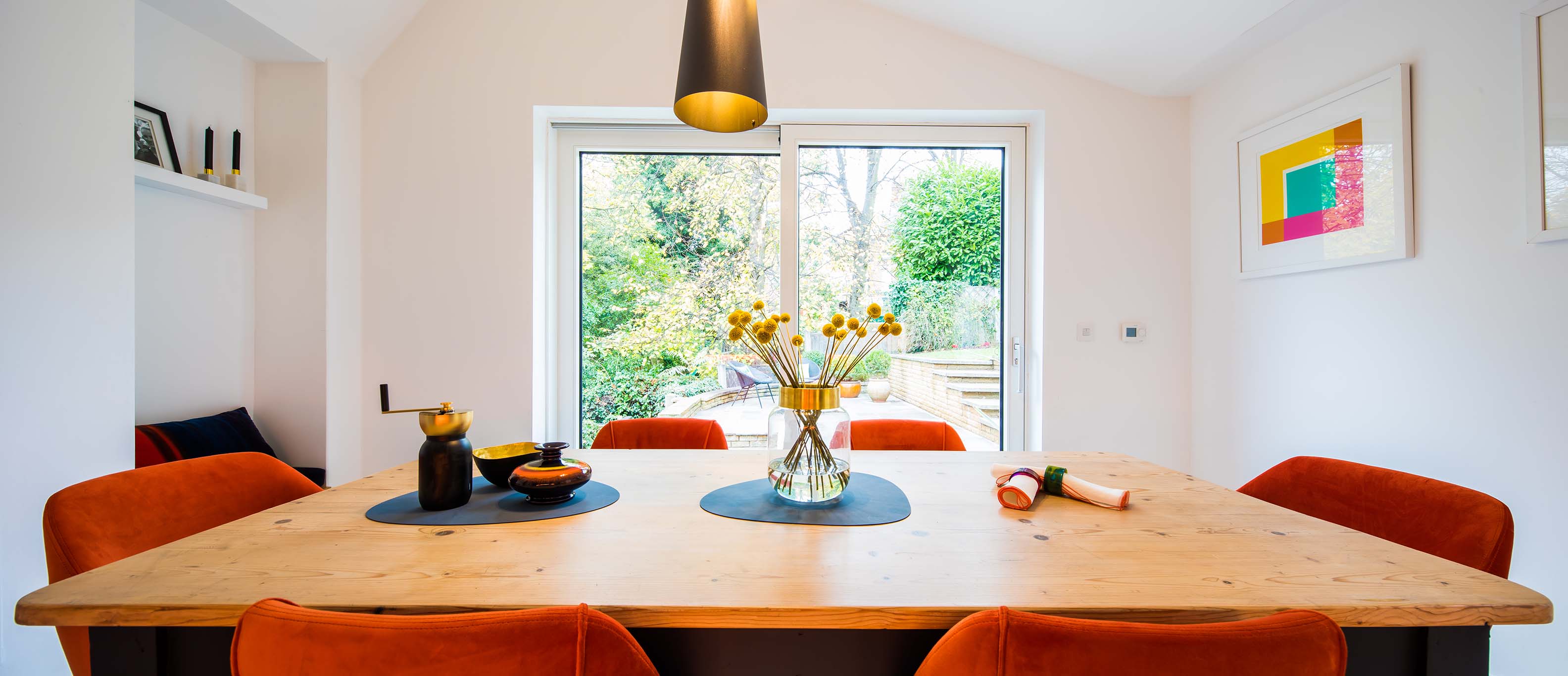 Dining area with a wooden table and orange chairs, featuring a large low u value window with a view of greenery.