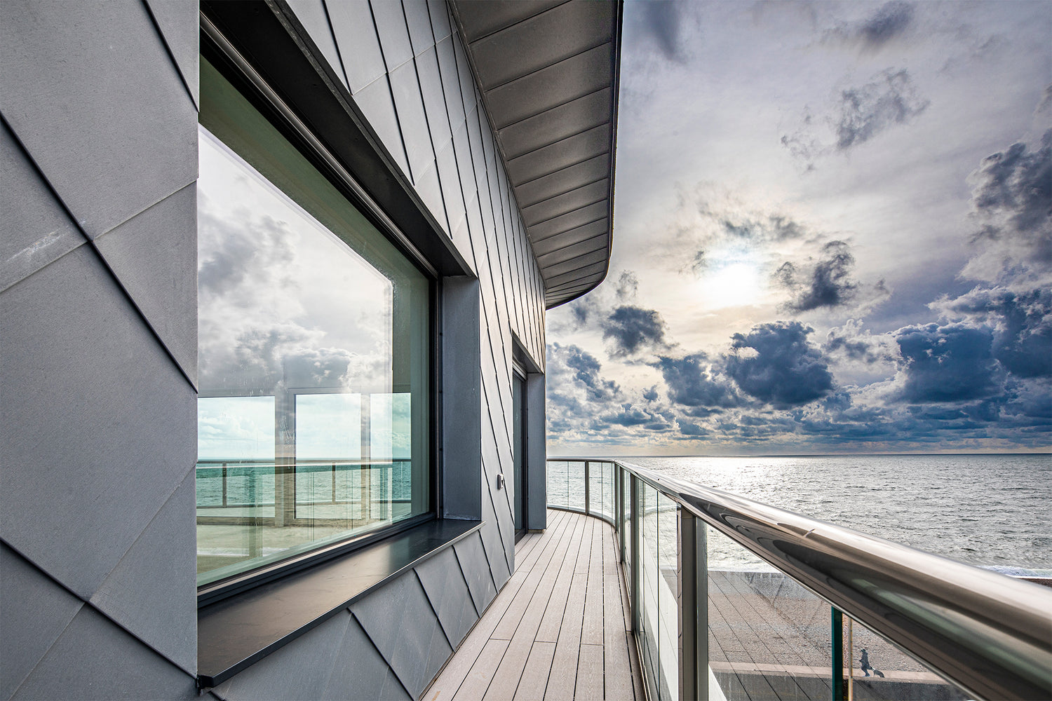 Modern building with triple-glazed window, a balcony overlooking a cloudy sky and water.