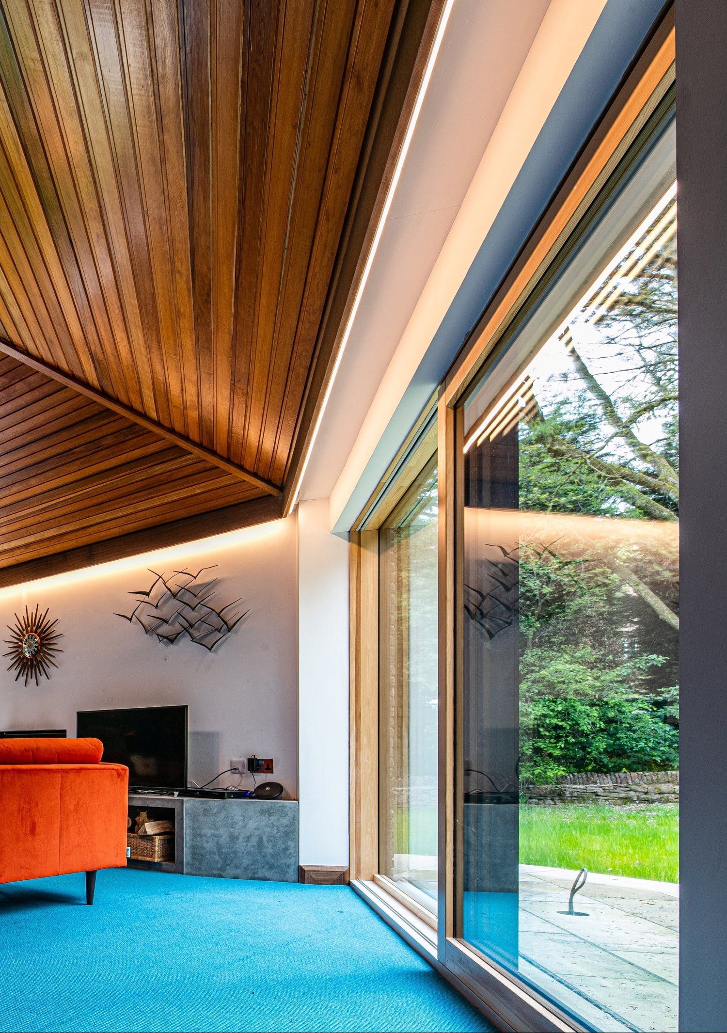 Passive house modern living room with orange sofa, large triple-glazed window, and wooden ceiling.