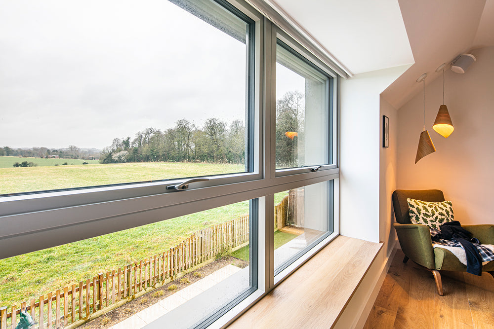 Modern room with large triple-glazed windows offering a view of a field, featuring a cozy corner with a chair and blanket.