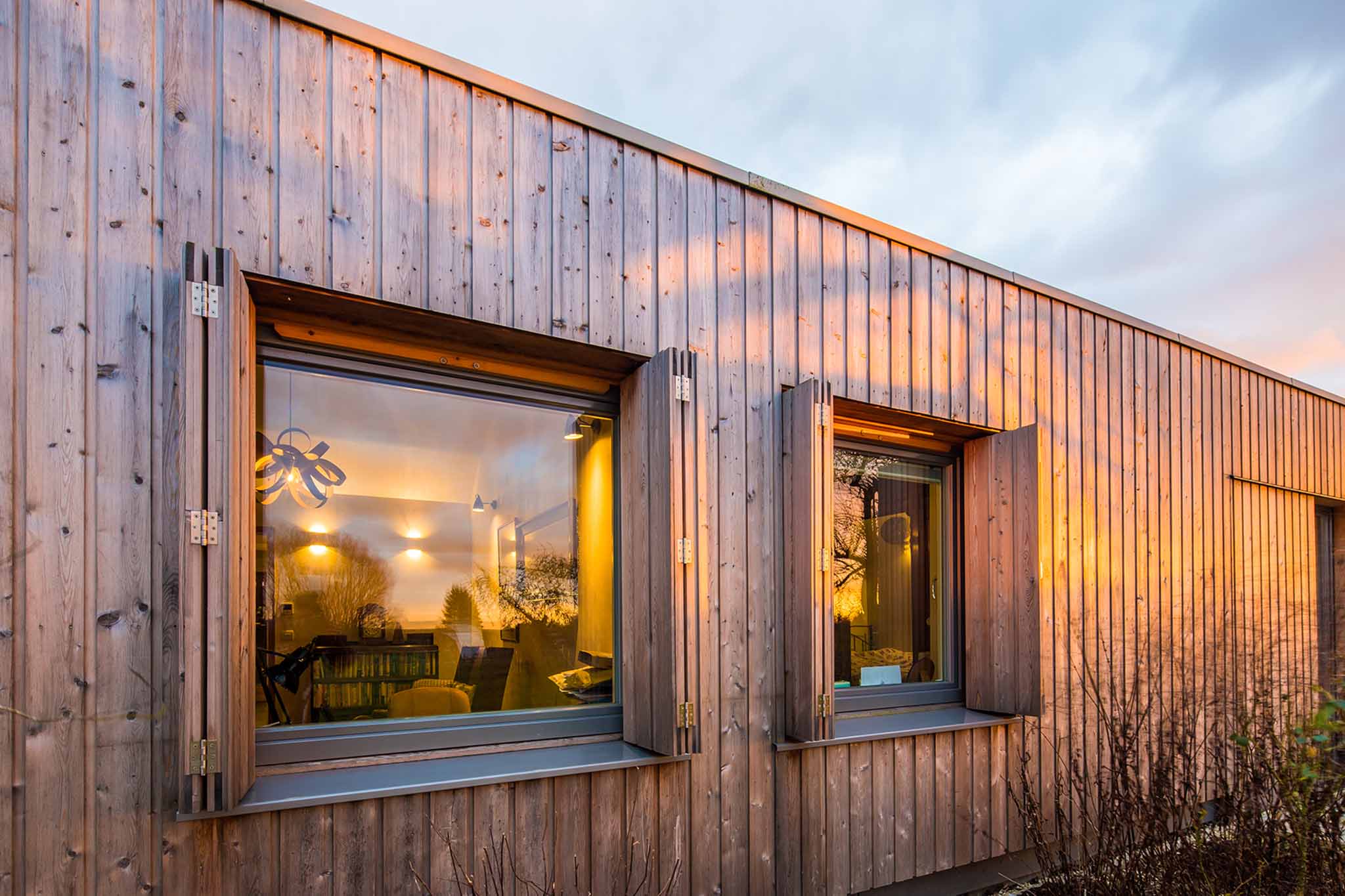 Wooden house exterior with two triple-glazed windows, reflecting indoor lights and furniture.