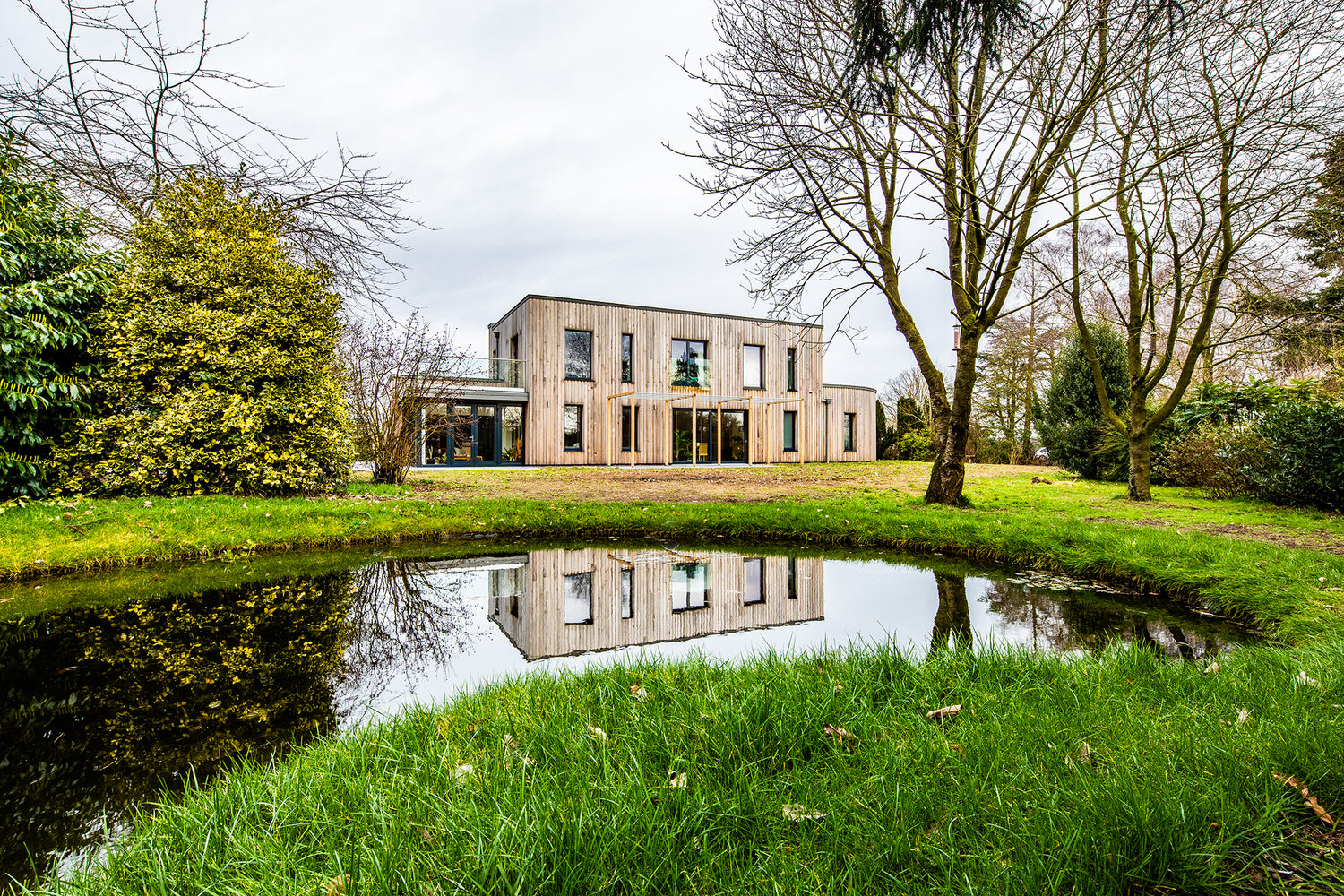 Modern, flat-roofed, timber clad home set across two levels with dark-framed composite windows and doors, seen from across a lake