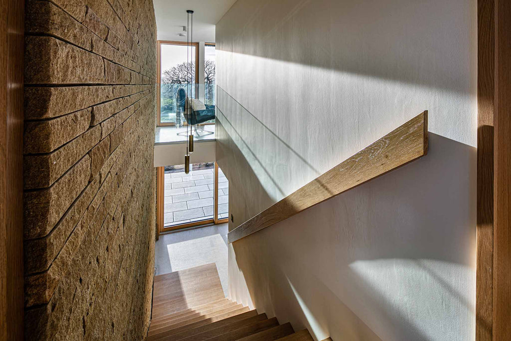 Modern staircase with triple-glazed windows, wooden handrail and stone wall in a well-lit room.