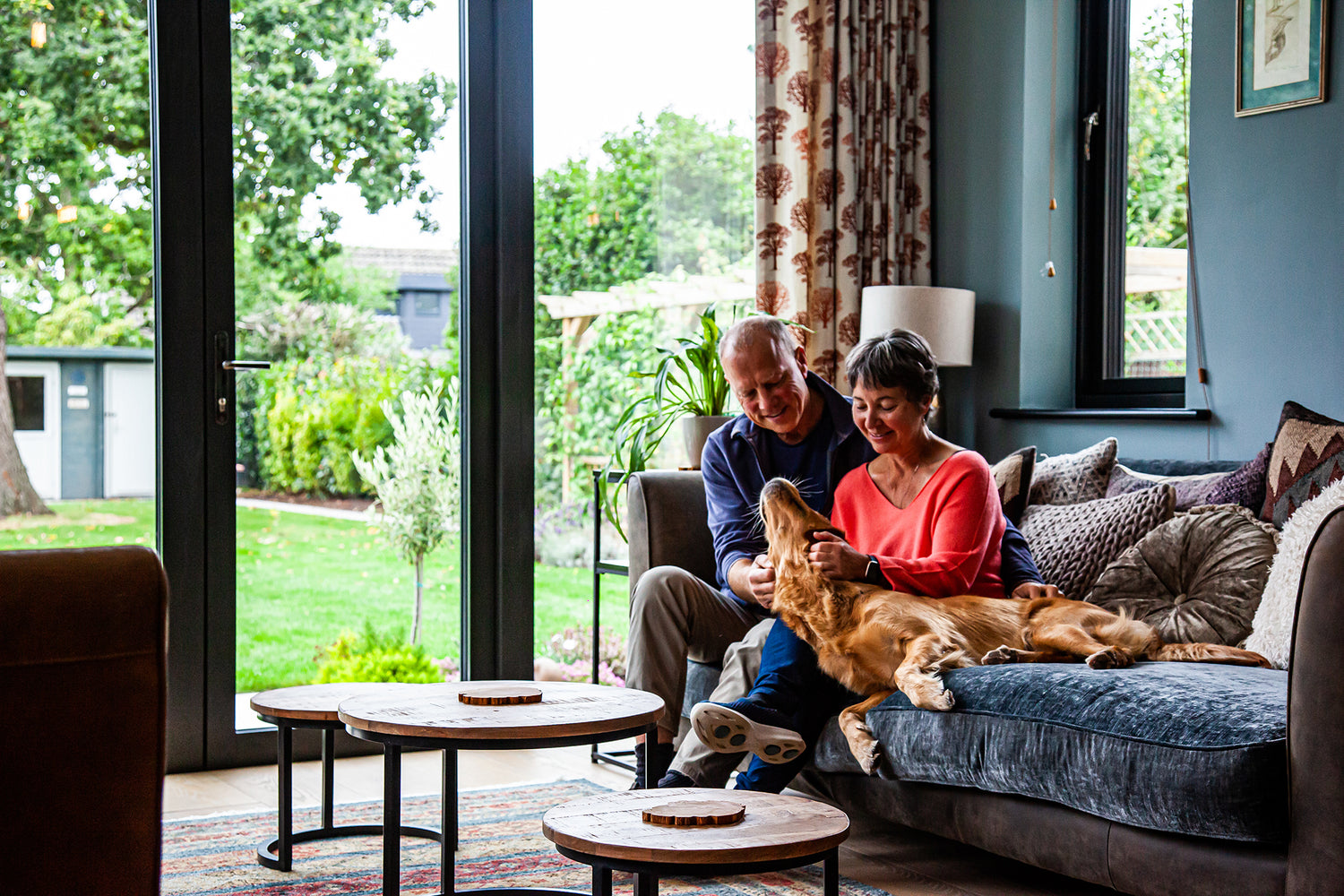 Man and woman sitting on a couch with a dog in a living room with large  triple-glazed windows.
