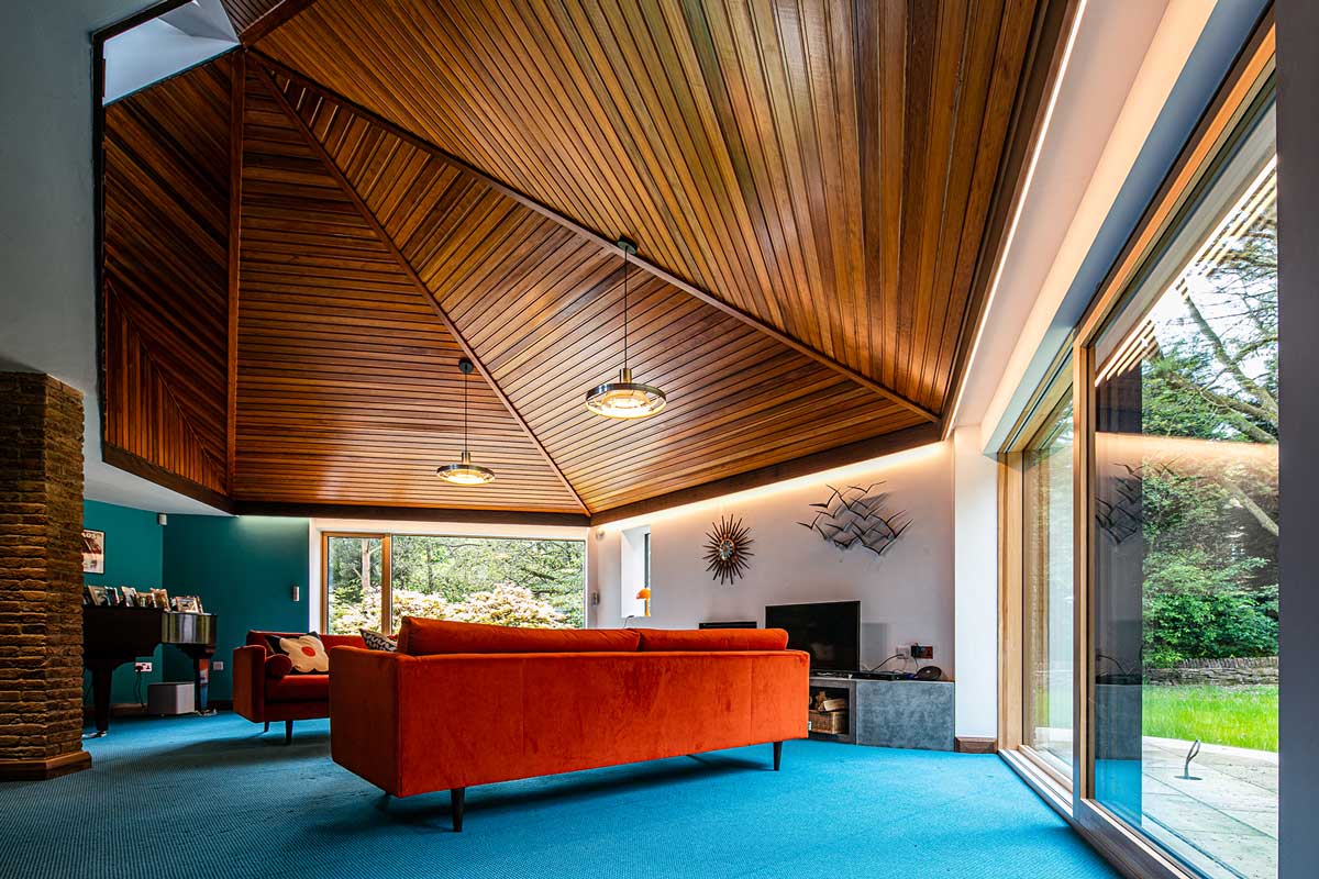 Image of a living room with a tall, timber-clad ceiling, blue carpets and large orange sofas. Timber sliding doors are on opposite sides of the room.