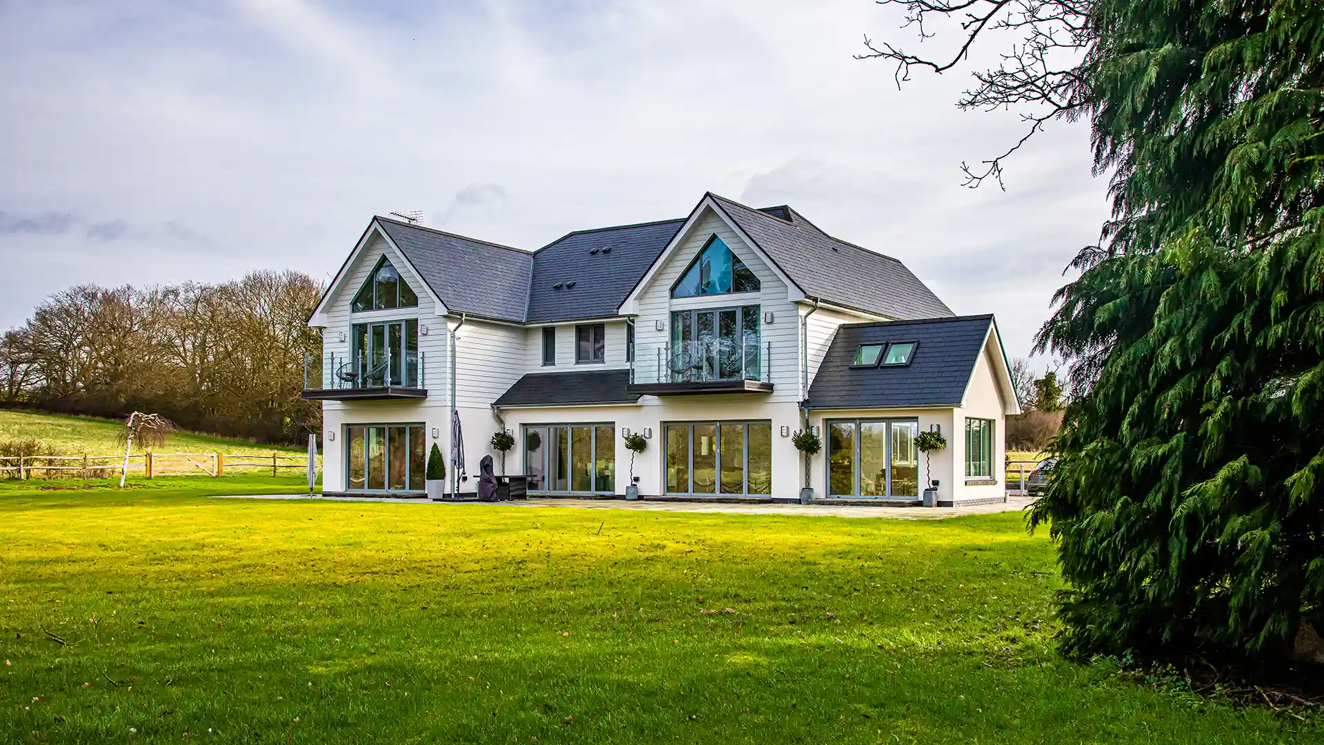 Image of a large, nordic-style new build home clad in white timber with grey aluminium-clad composite windows and bifold doors in a large open lawn space