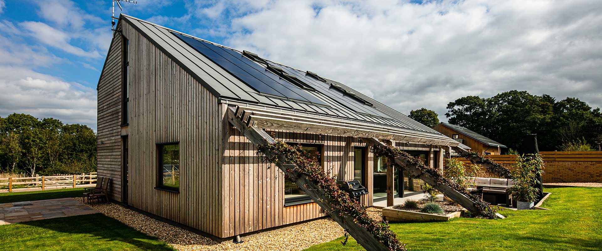 Image of a timber-clad new-build home with a slate grey roof, angled timber beams with climbing plants and black triple-glazed windows and doors