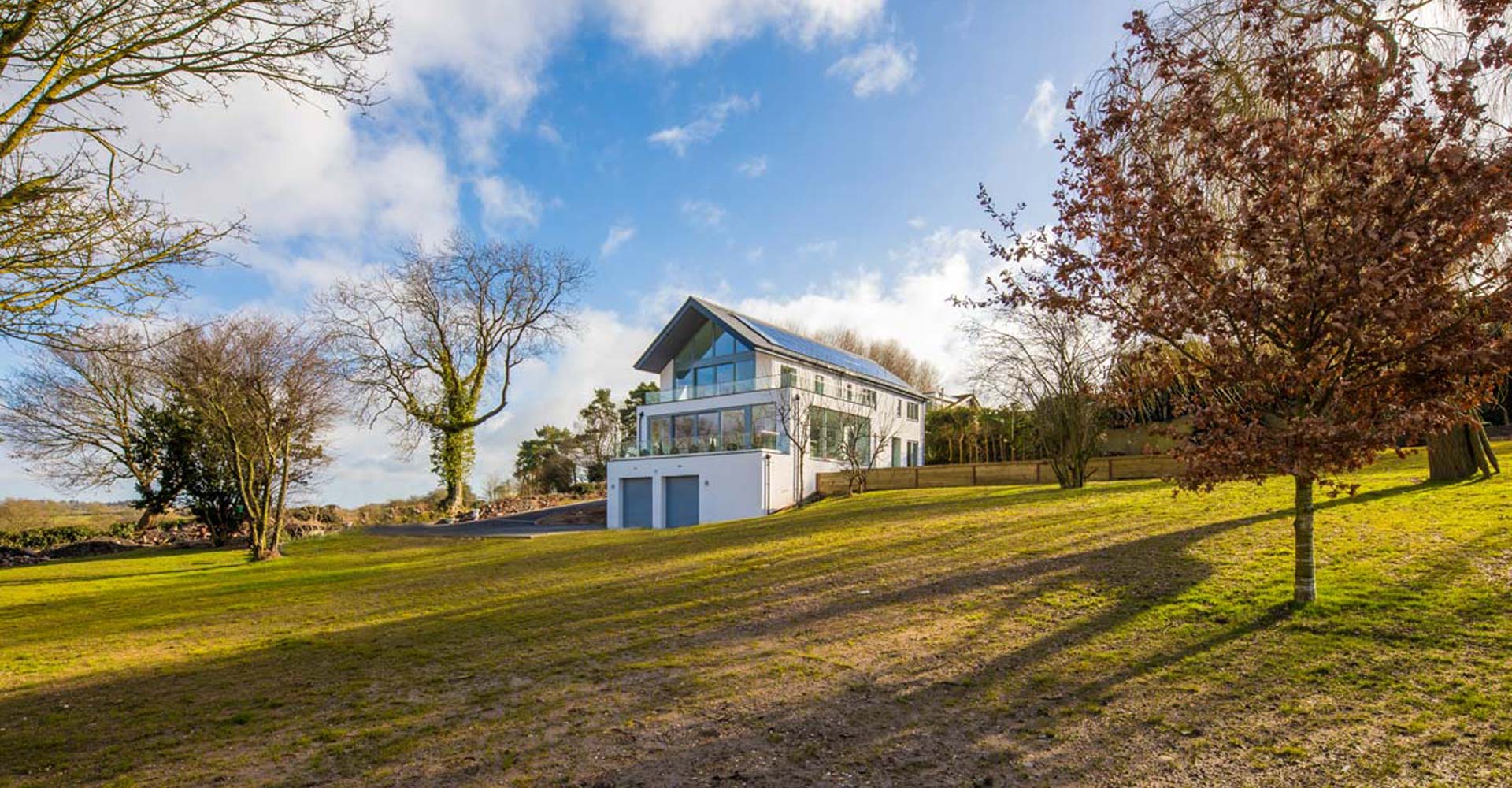 Image of a grey and white rendered modern home with glass balcony and a large gable of alu-clad windows and sliding doors set on a hill in a countryside setting