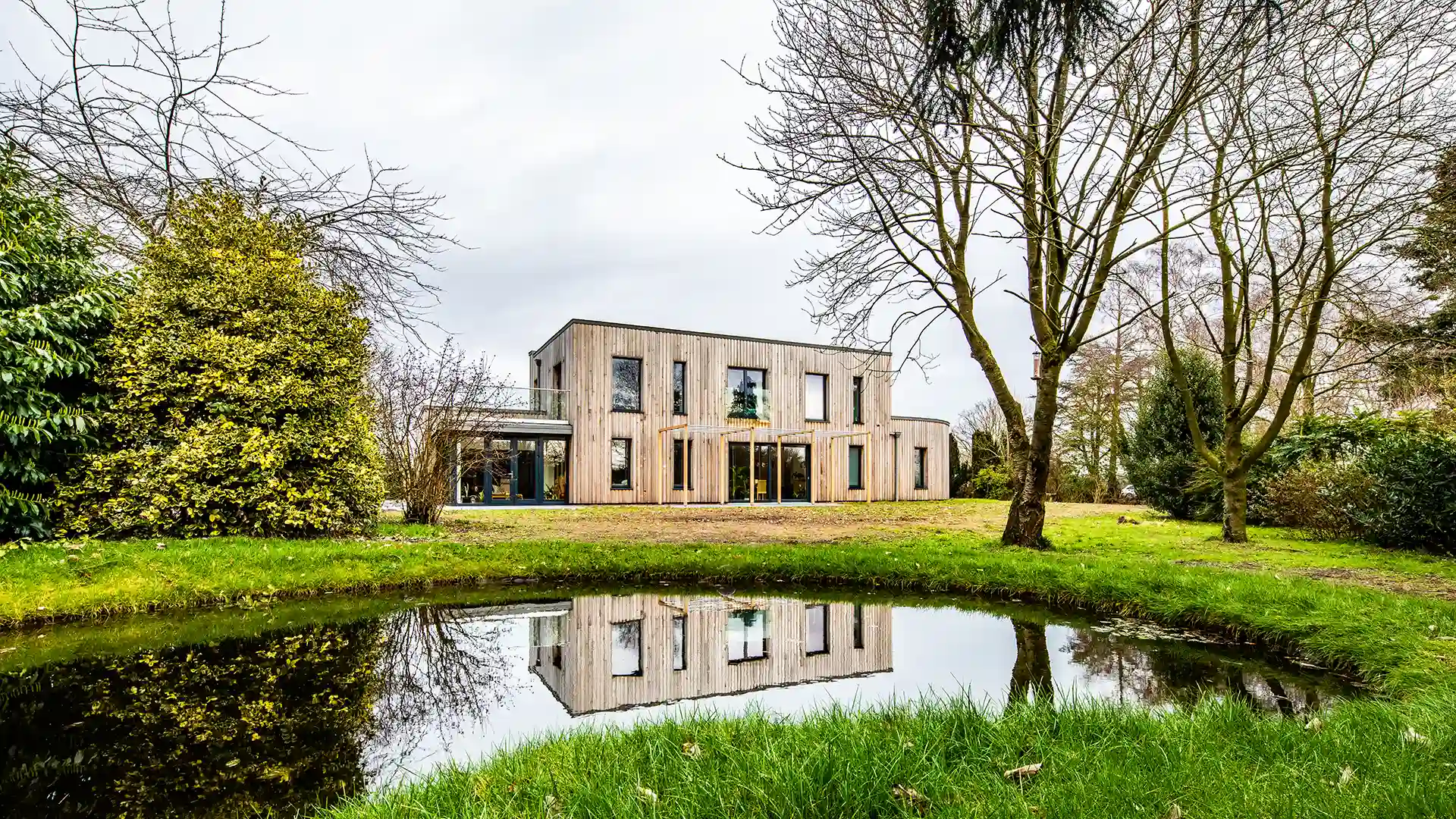 Image of a modern, flat-roofed, timber clad home set across two levels with dark-framed composite windows and doors taken from across a lake near the home